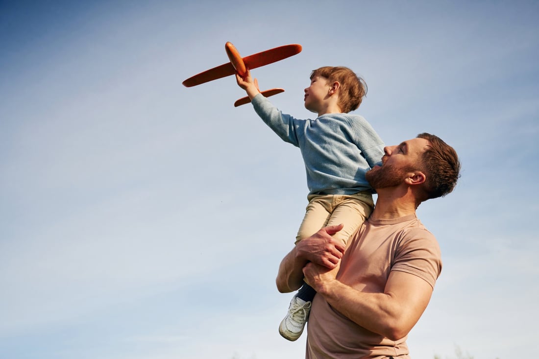 A father lifting his son, who is flying a toy plane, against a blue sky