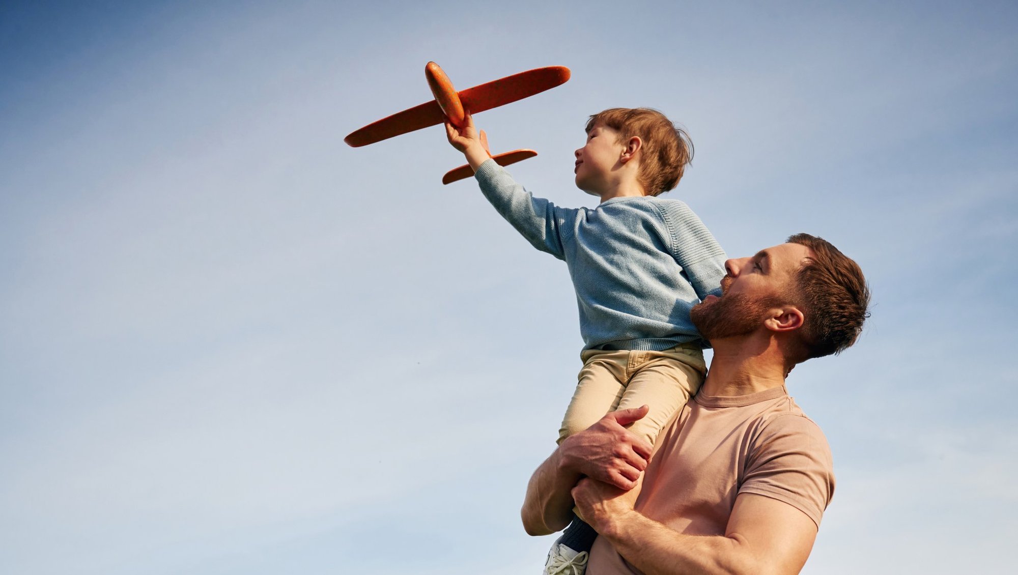 A father lifting his son, who is flying a toy plane, against a blue sky